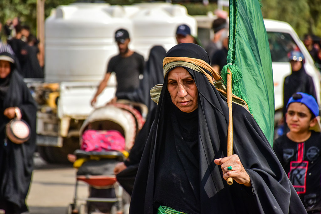 A woman pligrim wearing black carries a green flag over her shoulder an looks weary.