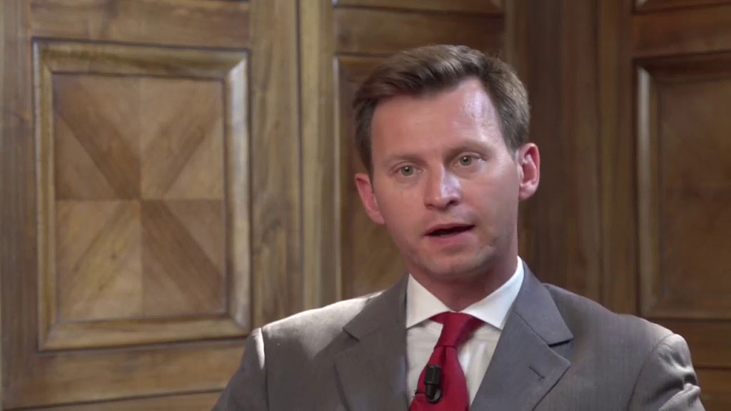 A man talks animatedly looking at the camera while sitting against a wood panelled wall.