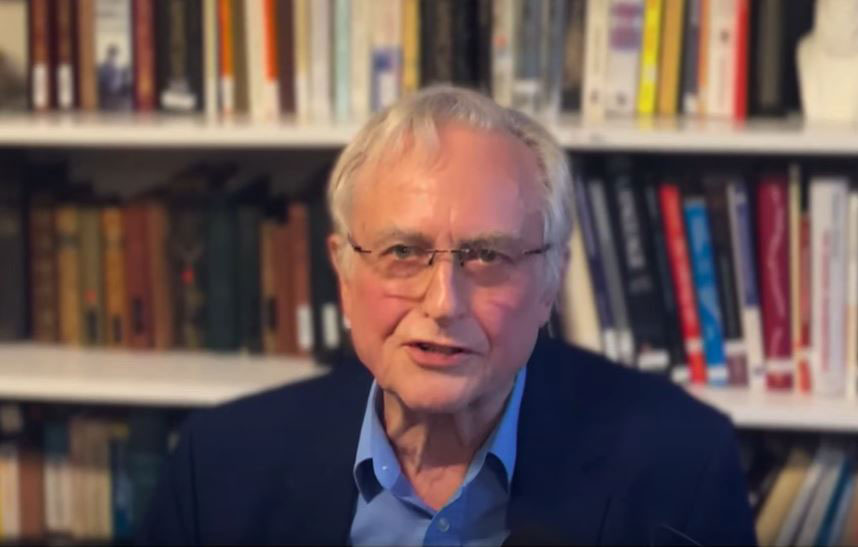 A man sits and speaks, against a background of a bookcase.