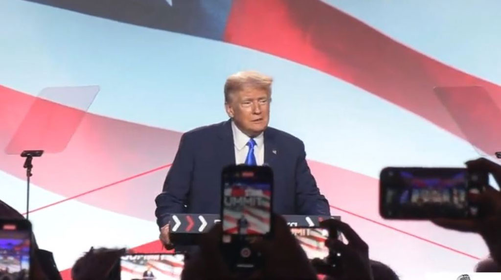 Donld Trump speaks against a US flag backdrop while the audience hold up phones.