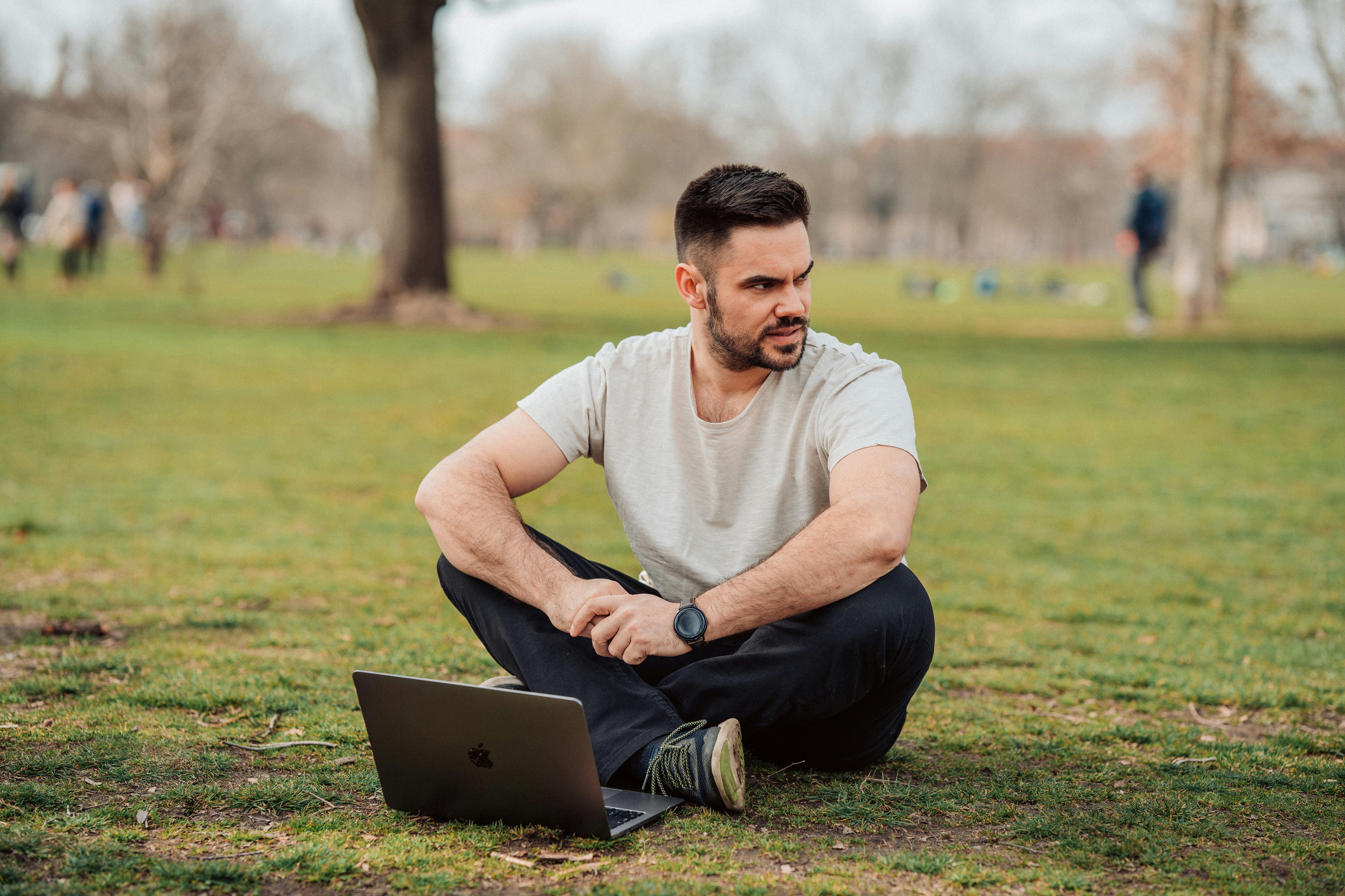 A man sits cross legged in a park with a laptop on the grass in front of him. He looks to one side.