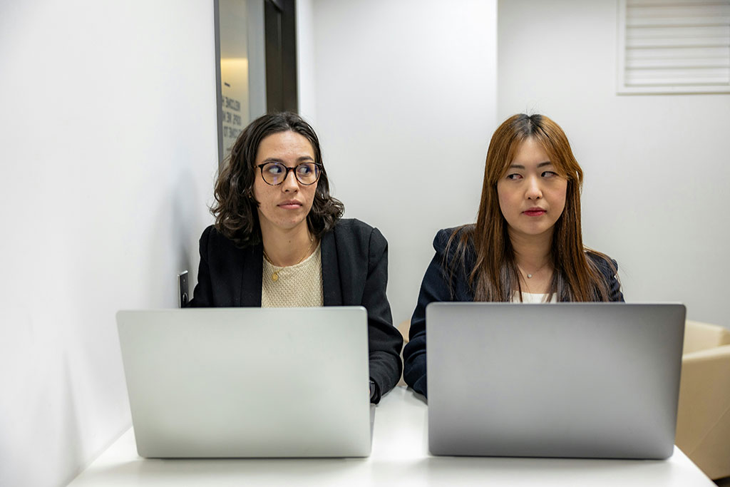 Two people sit beside each other working on laptops, One looks askance to the other who frowns.