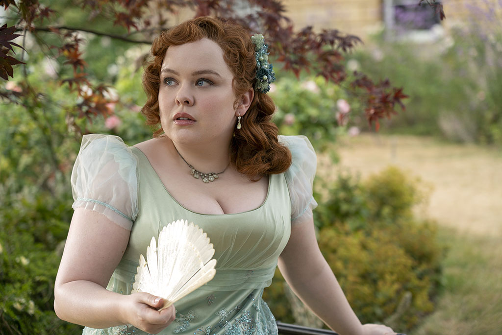 A young lady in Regency dress, holds a fan while looking around a garden.