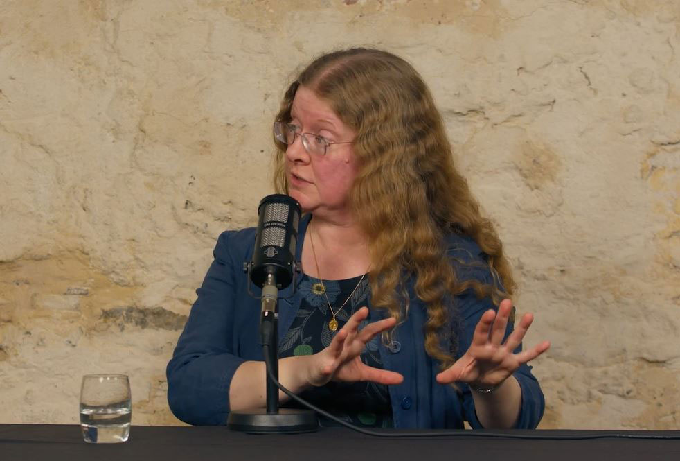 a woman seated in front a mic, looks to the left will gesturing both hands to the right.