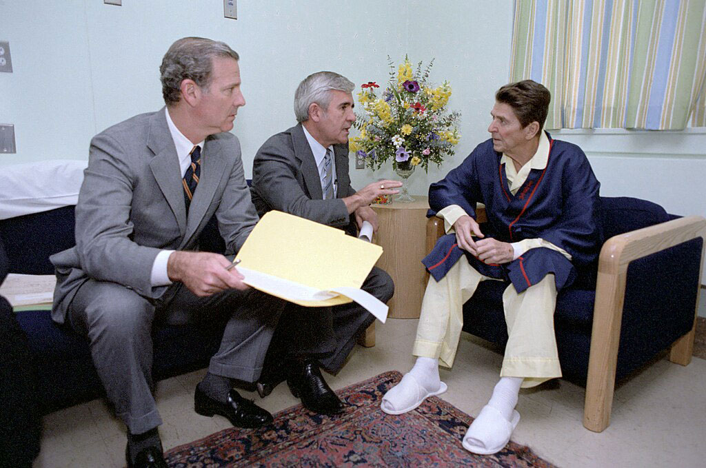 Two men in seats adress a pyjamap-clad Ronald Regan.
