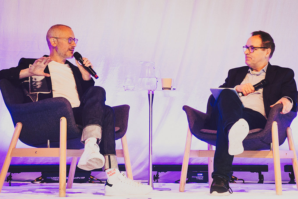 Two people sit on a stage in a relaxed manner, having a converation.