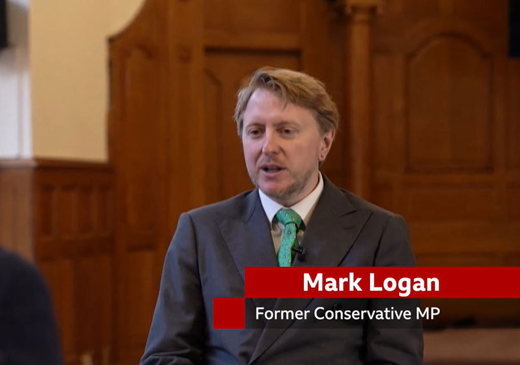 A man sits in a TV interview, below him is a captions stating 'Mark Lognan. Former Conservative MP