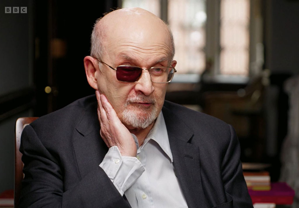 A man sits being interviewed and holds a hand to the side of his face, one lens of his glasses is tinted black.