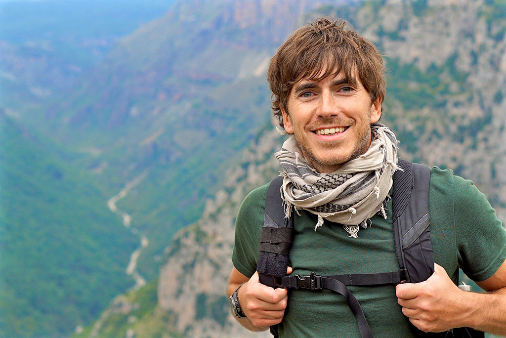 An enthusiastic hiker stands in front of a view down a valley, smiling and holding his backpack straps.