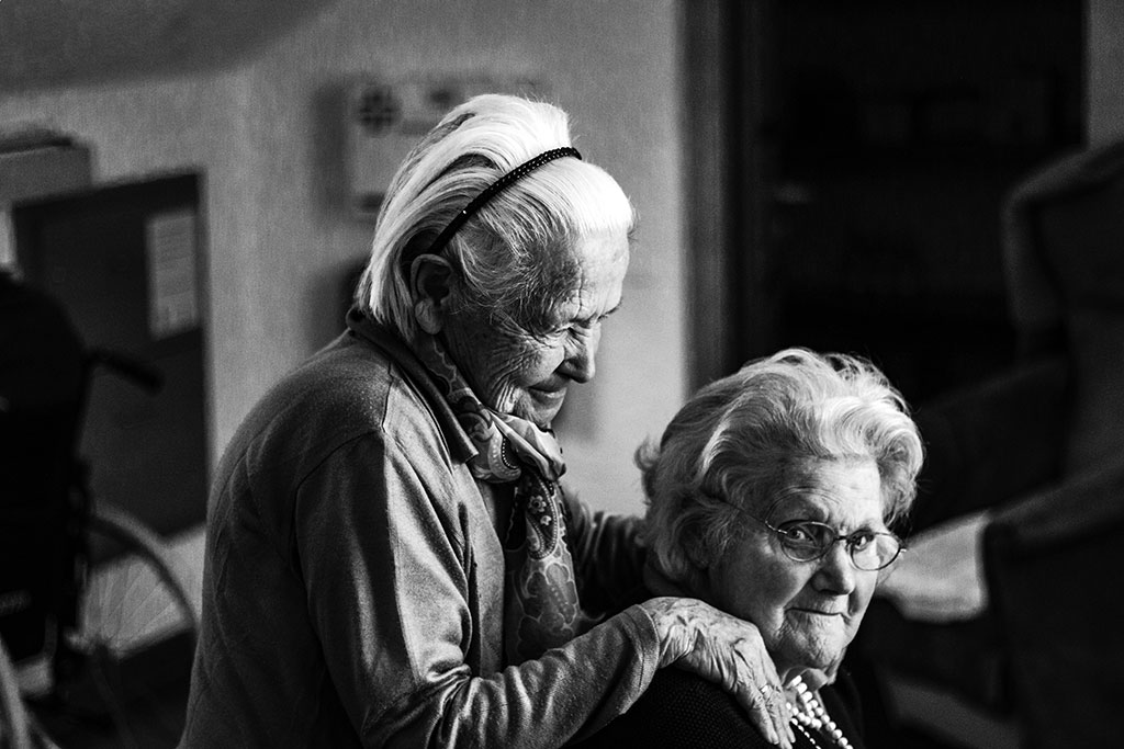 A elderly women in a care home stands and places her hands on the shoulders of a seated woman.