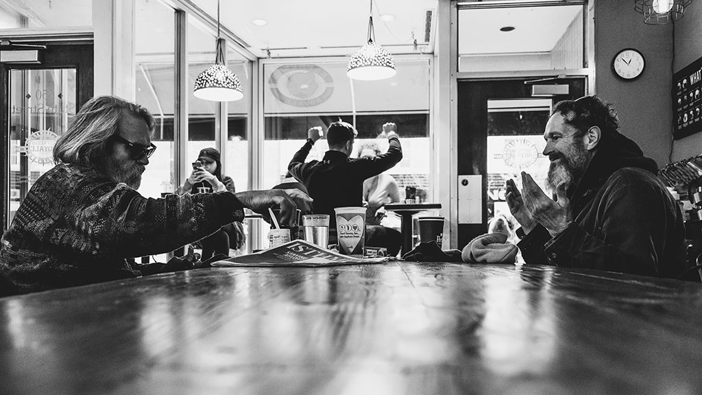 In a coffee shop, people sit around a table, one holds their hands up in surprise. 