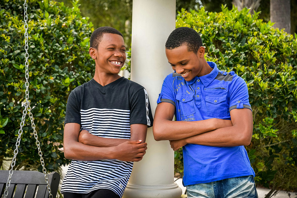 Two teenager lean against a rail, arms crossed, and laugh together.