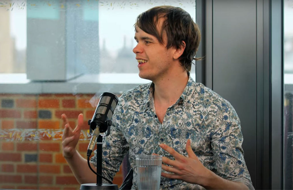 A podcast guest speaks in front of a mic and raises their hands to gesture.