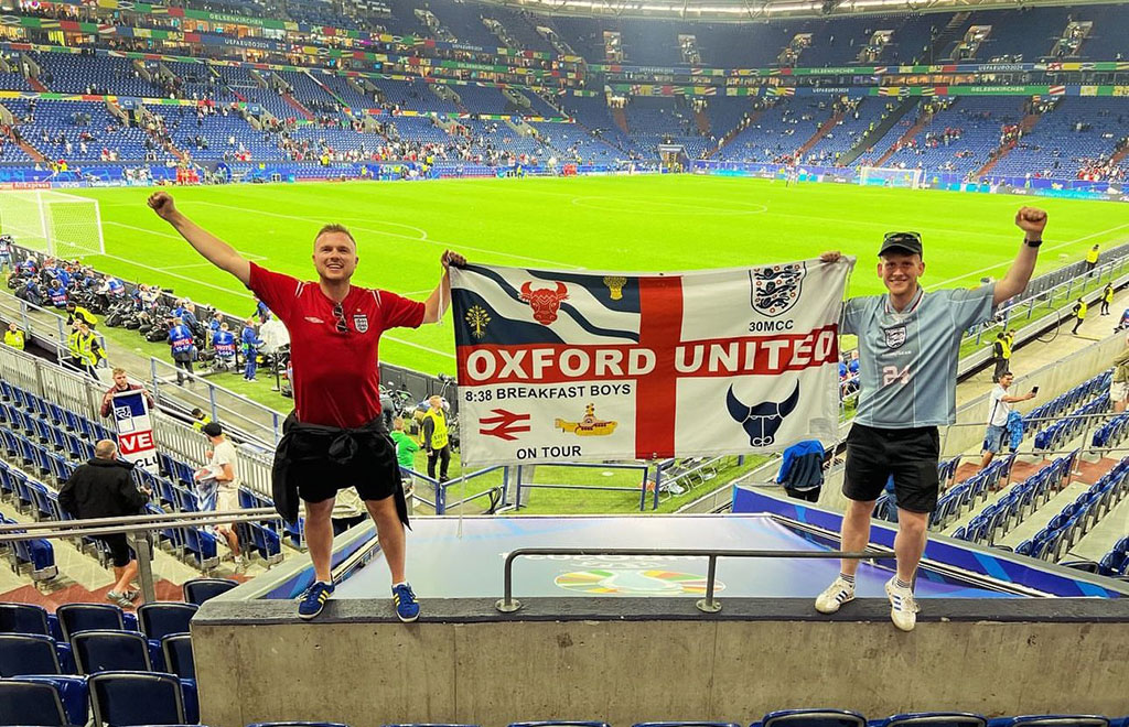 Two England fans stand in stadium seeting holding up their flag.