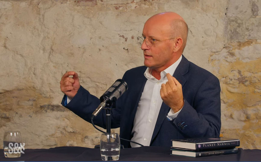 A man sits and speaks into a microphone and gestures with raised hands.