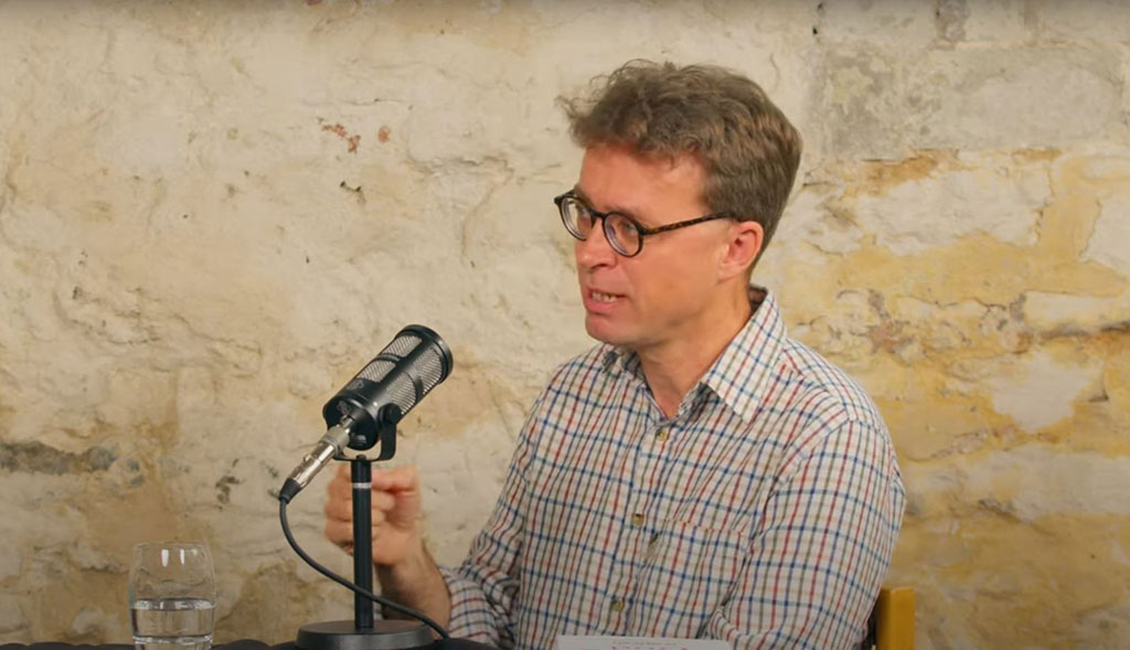 A podcast guest speaks in front of a mic and raises their hands to gesture.