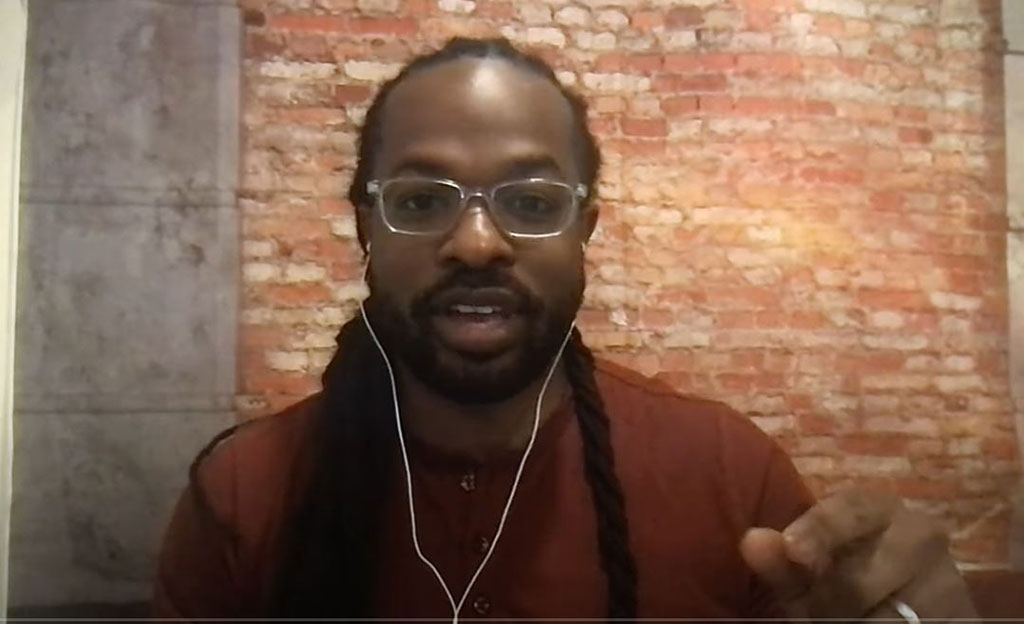 A man wearing white earphones talks to a camera and gestures with one hand.