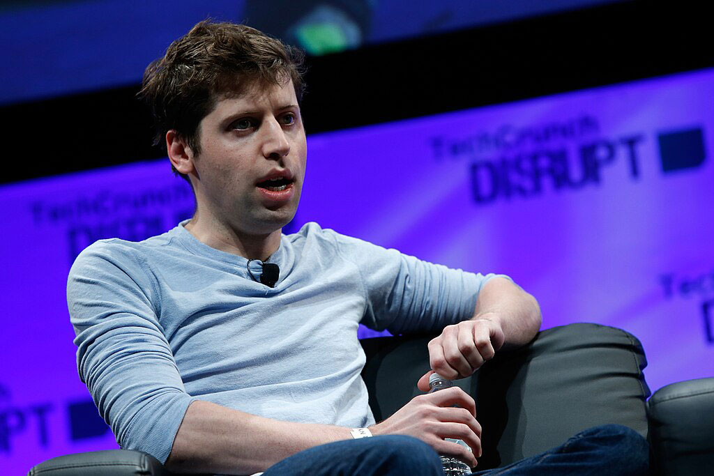 On a conference stage, a seated speaker leans back and opines