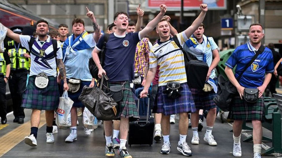 Scottish football fans wearing kilts march down a street singing and waving their arms alogt.