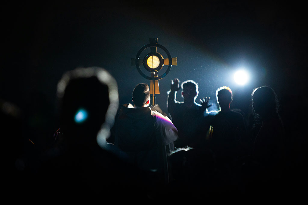 A cross held aloft is illumminated by a shaft of light that also reveals hands raised in priase.