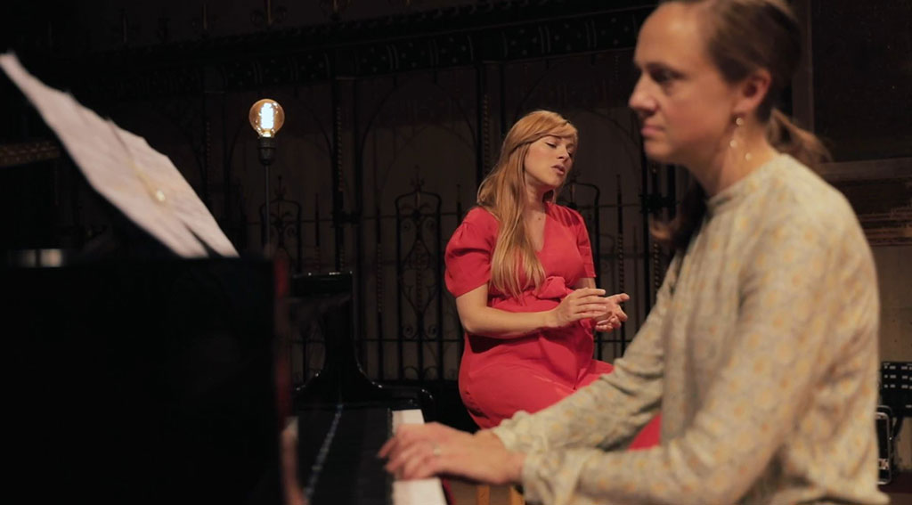A pianist plays in the foreground and a seated singer gestures with eyes closed behind 