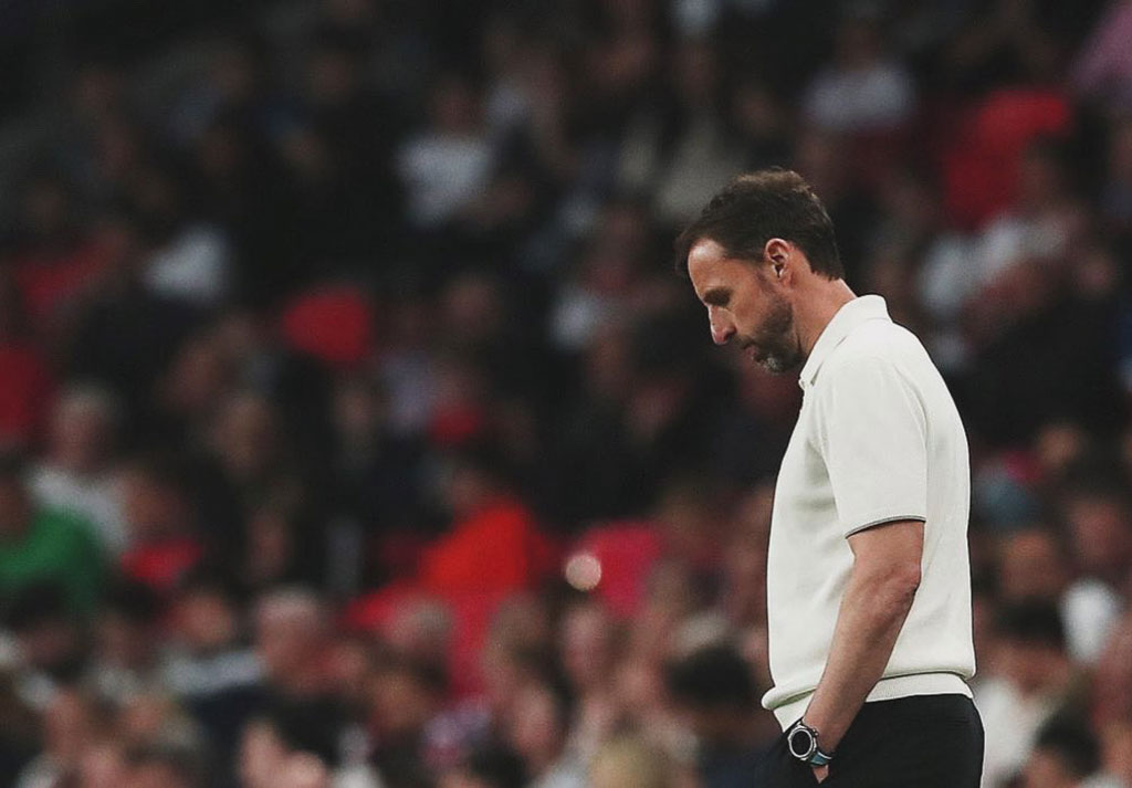 A dejected looking football manager ponders his feet while standing beside a pitch.