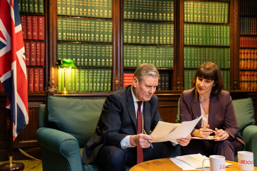 Kier Starmer sits on a sofa, leaning forward and holding papers he is reading. Rachel Reaves sits and looks on.