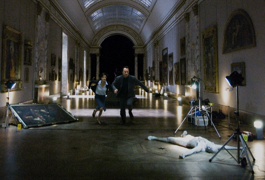 Two people run though a darkened art gallery towards a body lying amongst photography paraphanalia