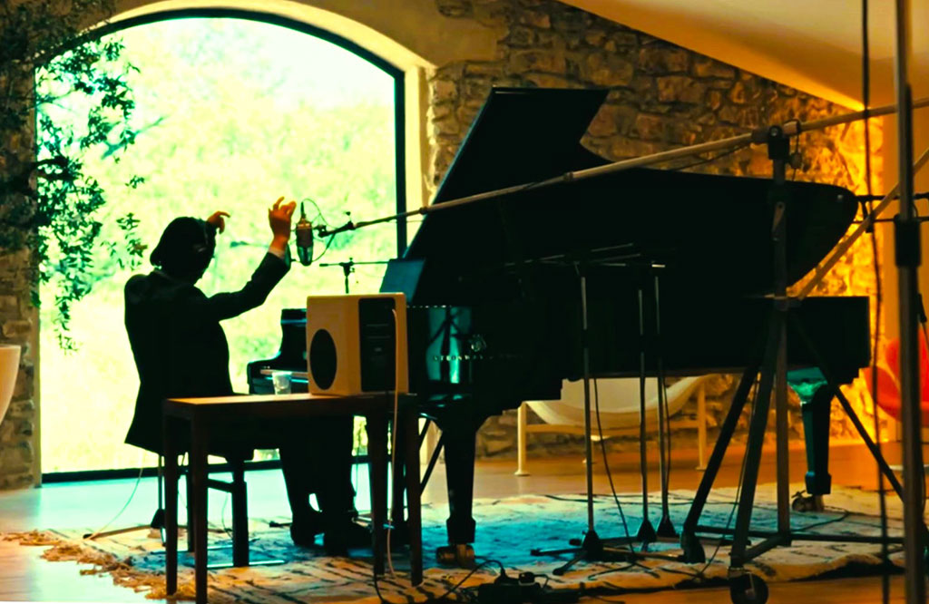 A pianist raises his arms while sitting at a grand piano amid recording equipment.