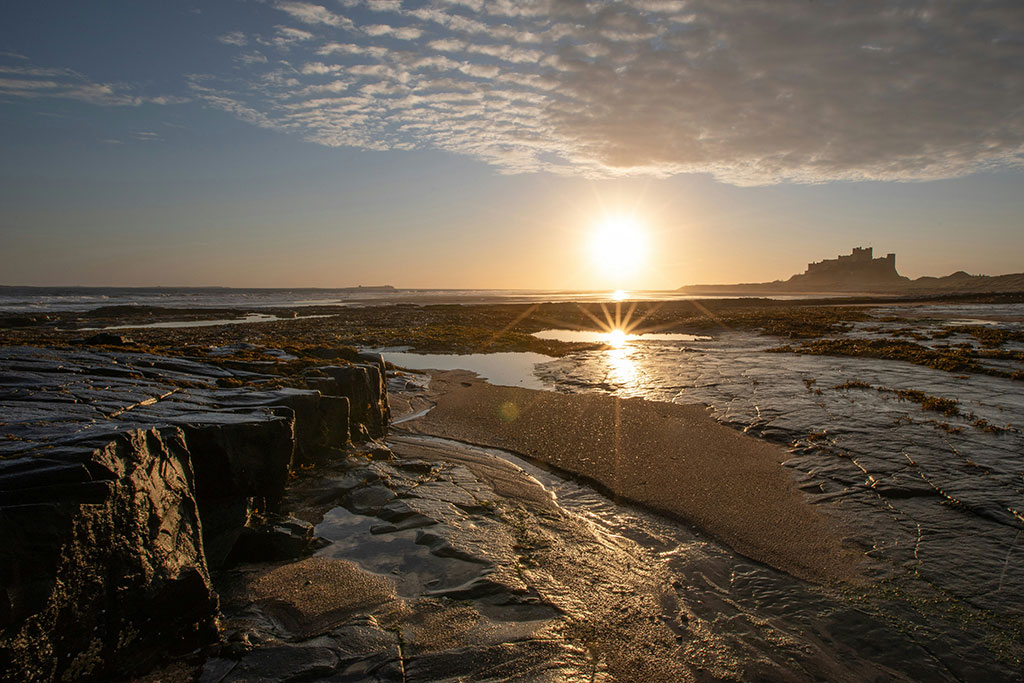 A rockpool on a beach reflects the sun, a castle stands beyond the sand dunes
