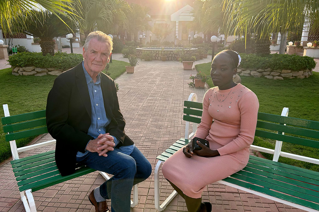 A TV interviewer sits across from a young woman in an outdoor setting