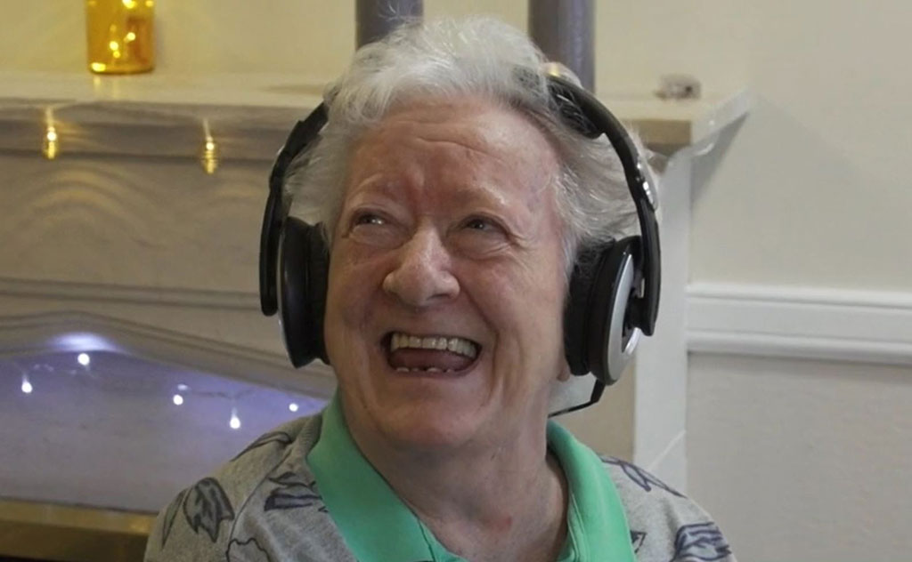 An elderly woman wearing headphone looks up and to the side with a big smile.