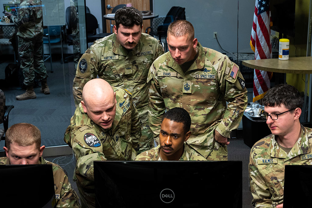 Military personnel, wearing camoflague uniform crowd round a computer monitor.