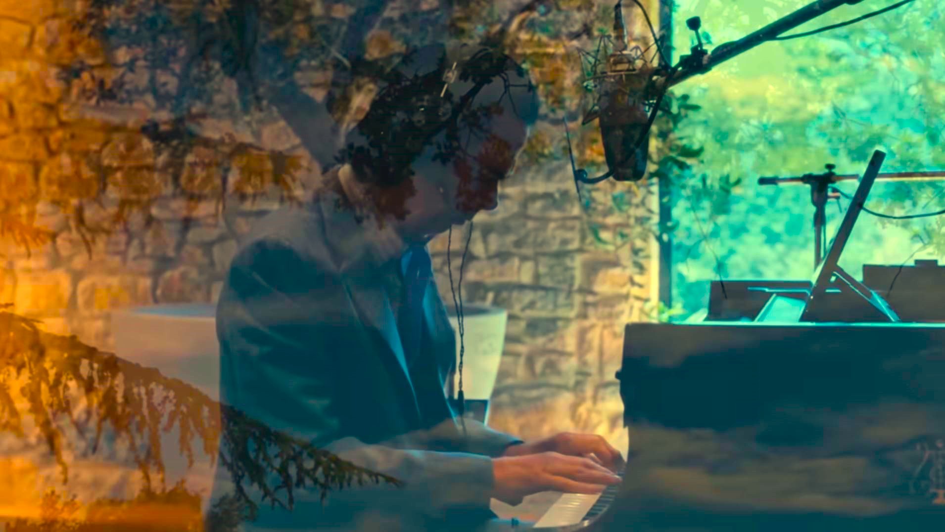 A pianist raises his arms while sitting at a grand piano amid recording equipment.