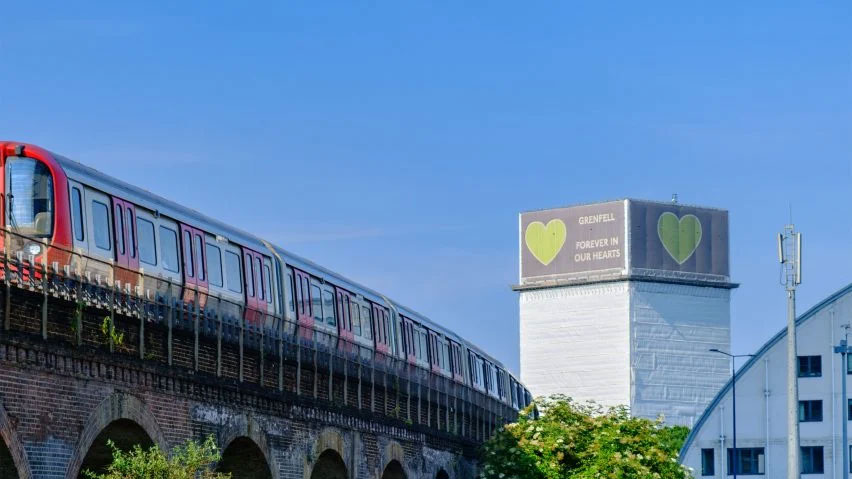 A tube train runs on a raised track, in the distance is a tower block wrapped in white material with a green heart on it.