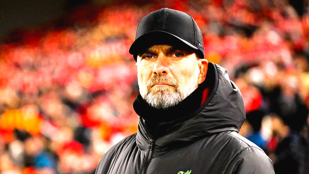 A stern looking  football manager stares hard, with a stand of supporters behind him.