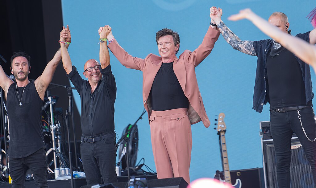 On a music festival stage, a popstar in a pink stages holds raised hands with his band.