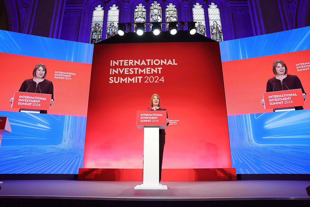 A woman stands behind a lectern against a blue and red backdrop.