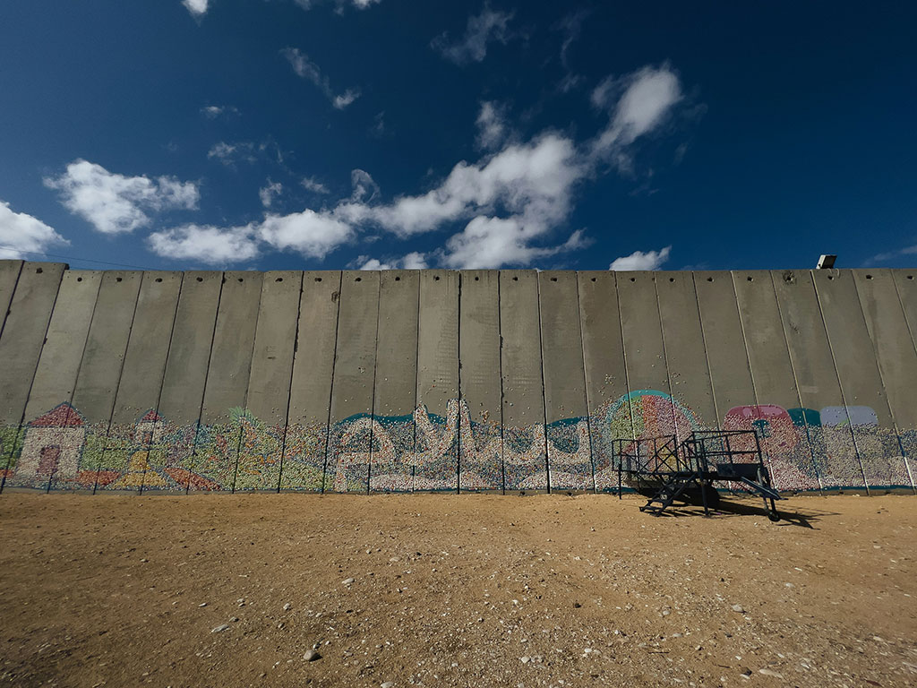 A graffitied concrete border wall stands below a blue sky and dusty ground
