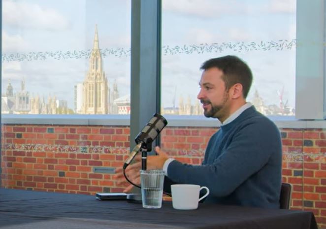 A podcast guest sits at a table talking with gesturing with their hands.