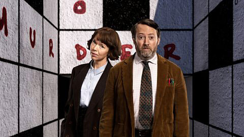 Two TV characters, a man and a woman, stand in front of a crossword cover walls of a room. 