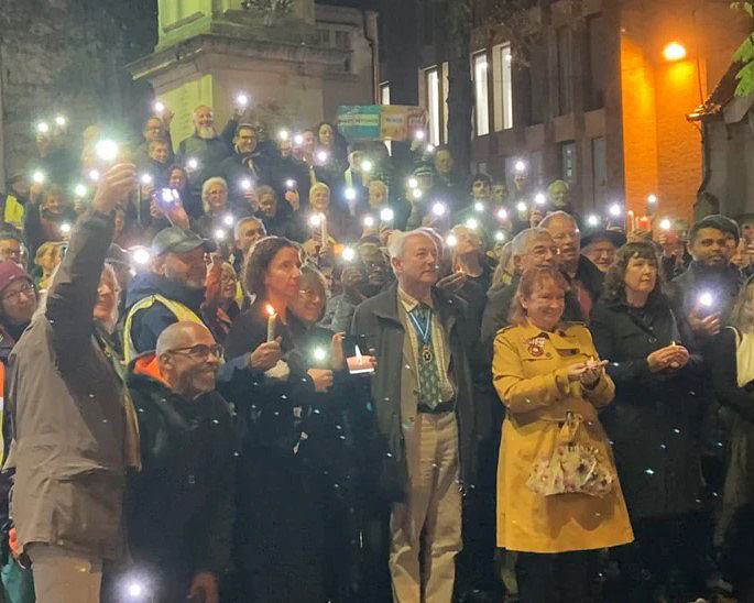 An outdoor vigil is lit by people holding up mobile phone lights.