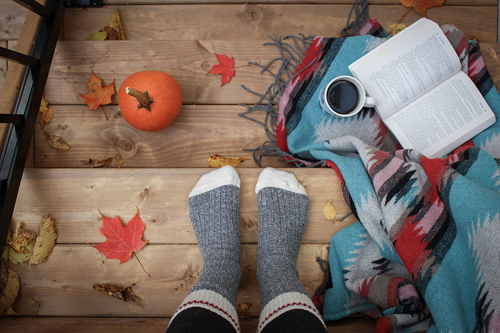 A set of be-socked feat rest on a leaf strewn step beside a book and a cup of coffee.