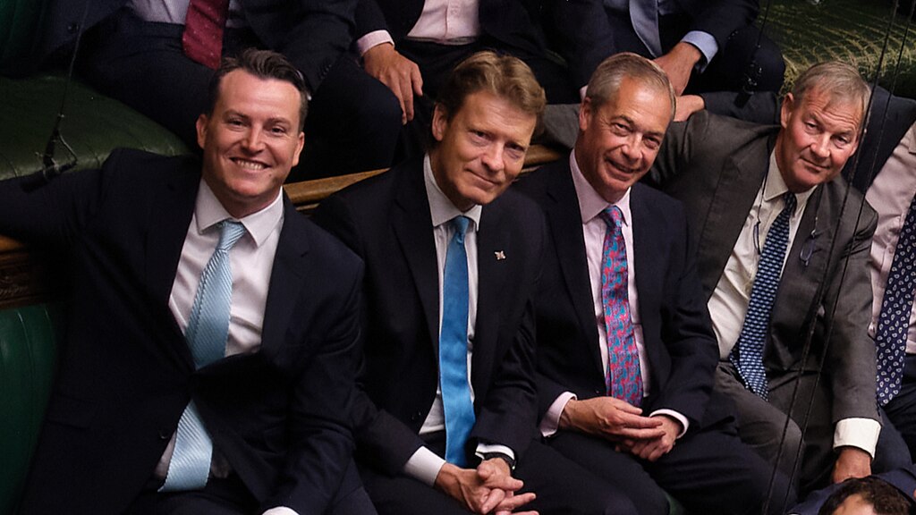 Four men in suits, sit next to each other smiling, in the House of Commons.