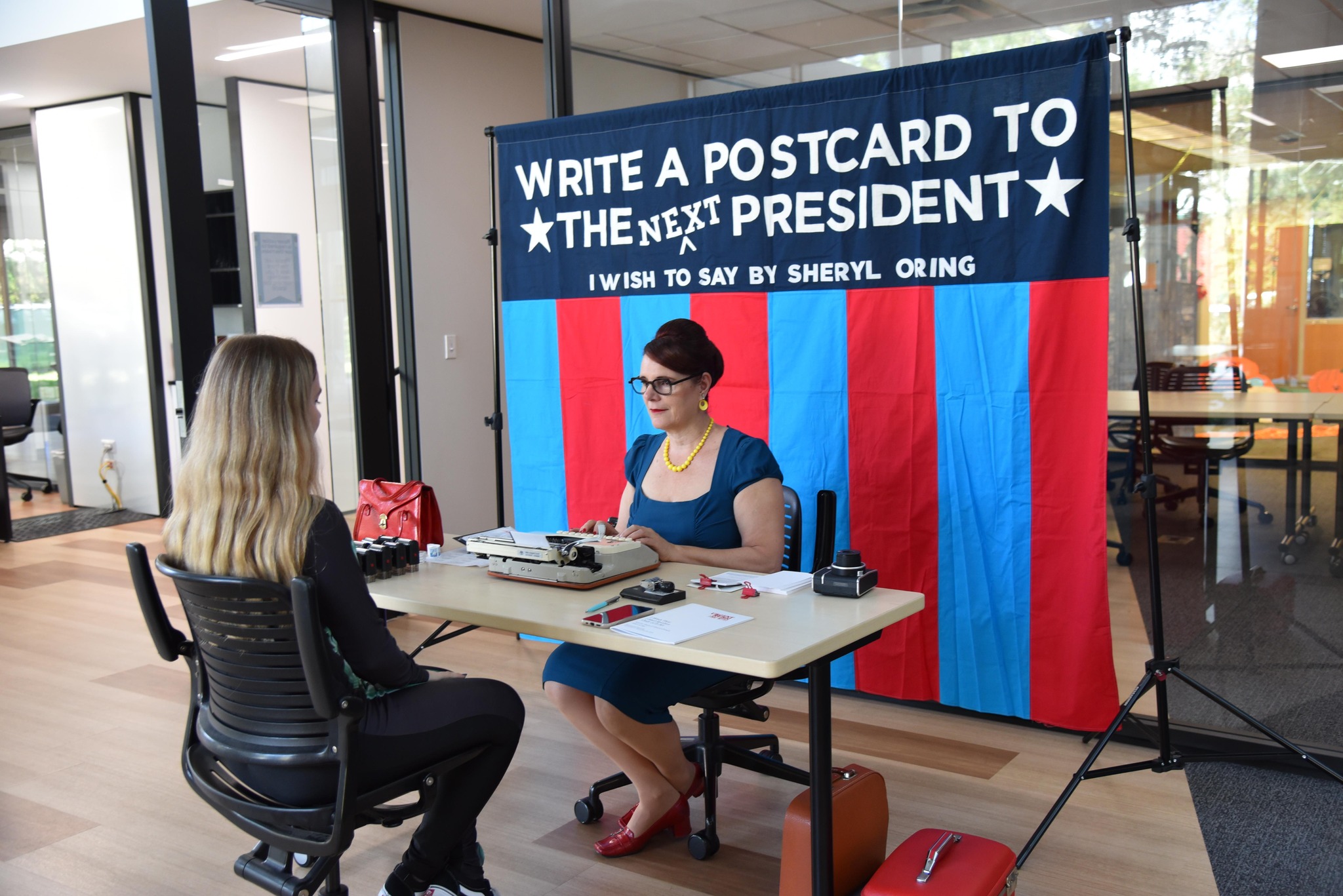 Two people sit at the table, one dictates as the other types. Behind a banner reads: write a postcard to the next president.i