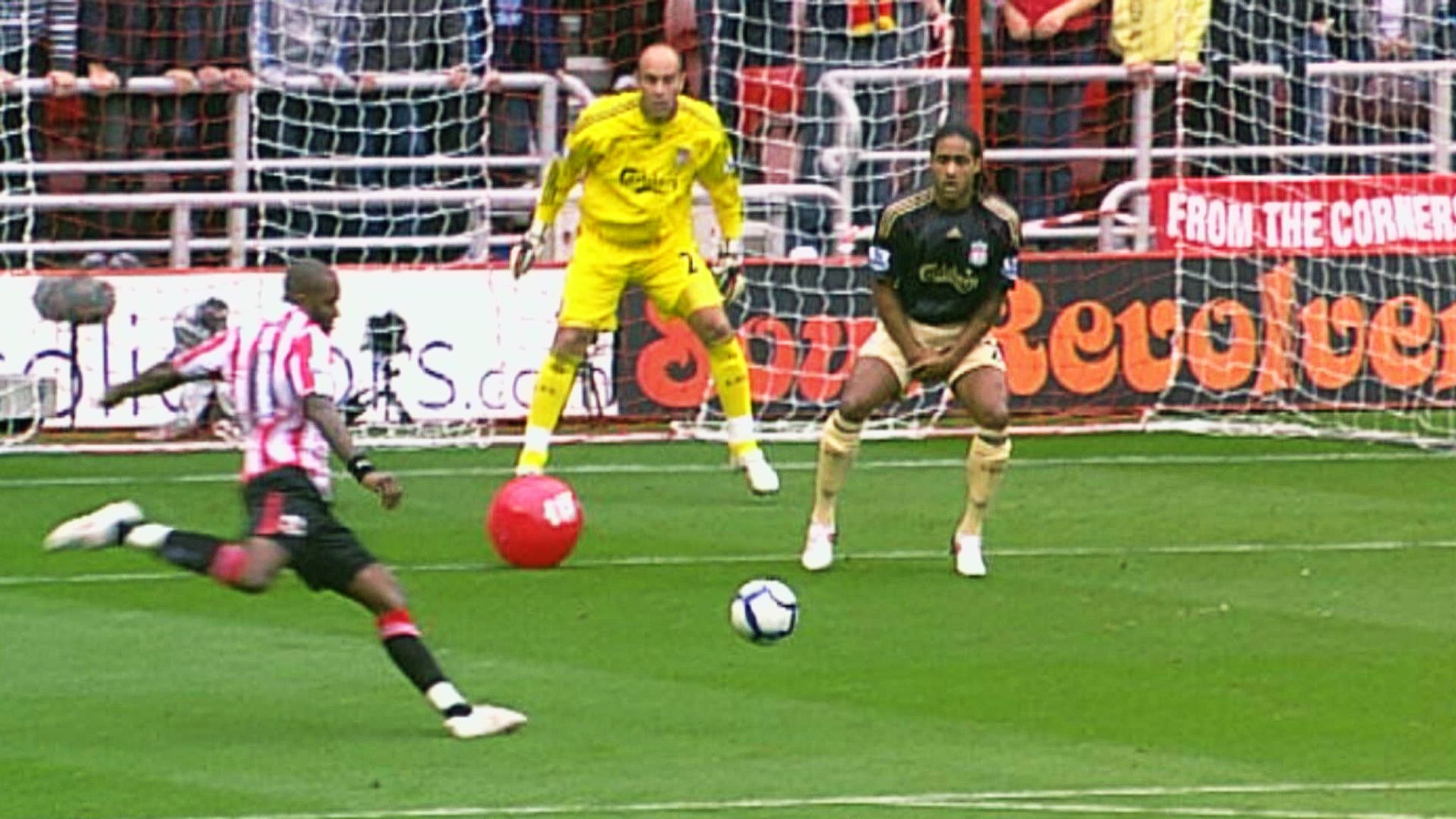 A striker is about kick a football towards a goal, a red beach ball sits between him and the goalkeeper.