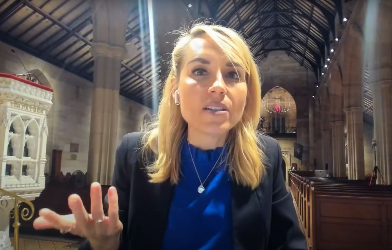 A woman sitting in an empty church talks to the camera and gestures with one hand.