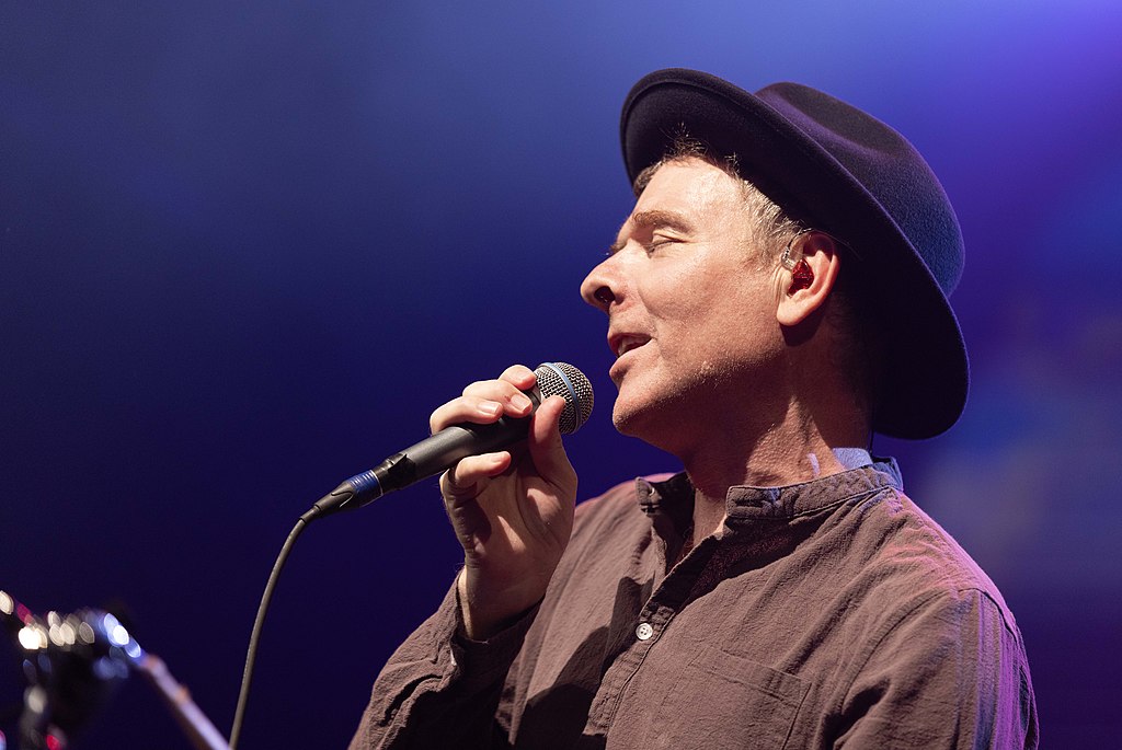 A singer, wearing a hat, pulls his head back holding a note, and a mic.