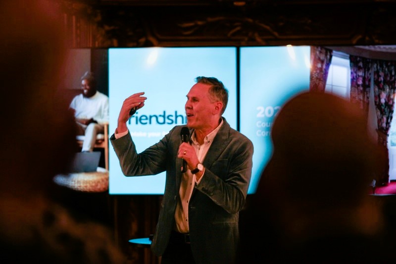A speaker, standing in front of a screen, beckons with one hand, holding a mic with the other.
