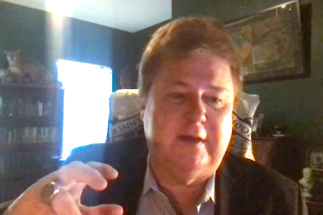 A podcast guest, on a screen, gestures with a raised hand.